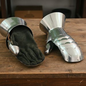 Pode incluir: Dois luvas de armadura medieval em metal polido. As luvas t&ecirc;m placas articuladas e s&atilde;o forradas com luvas verde-escuras. As luvas s&atilde;o exibidas em uma superf&iacute;cie de madeira. Artigo hist&oacute;rico.