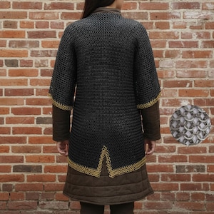May include: A black chainmail shirt with short sleeves and gold trim. It is worn over a brown padded gambeson. A close-up of the chainmail is in the lower right corner. The background is a brick wall.