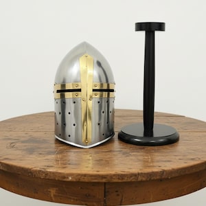 May include: A medieval-style helmet with a silver finish, brass accents, and a cross design. The helmet sits on a wooden table next to a black display stand. The helmet has small ventilation holes.