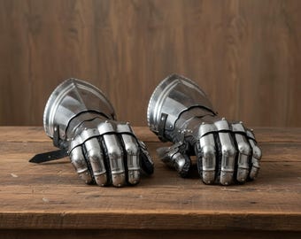 May include: A pair of medieval-style metal gauntlets, featuring articulated fingers and a polished, reflective surface. The gauntlets are made of silver-colored metal and rest on a wooden surface. The gauntlets have leather straps.