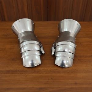 May include: Pair of polished metal gauntlets, likely for armor, with articulated fingers and wrist protection. The gauntlets are silver in color and appear to be made of multiple overlapping plates. The design suggests historical or fantasy themes.