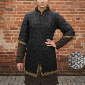 May include: Black chainmail shirt with gold trim on the sleeves and hem. The shirt has brown padded sleeves and a brown collar. A close-up of the chainmail is in the lower right corner. The background is a brick wall.