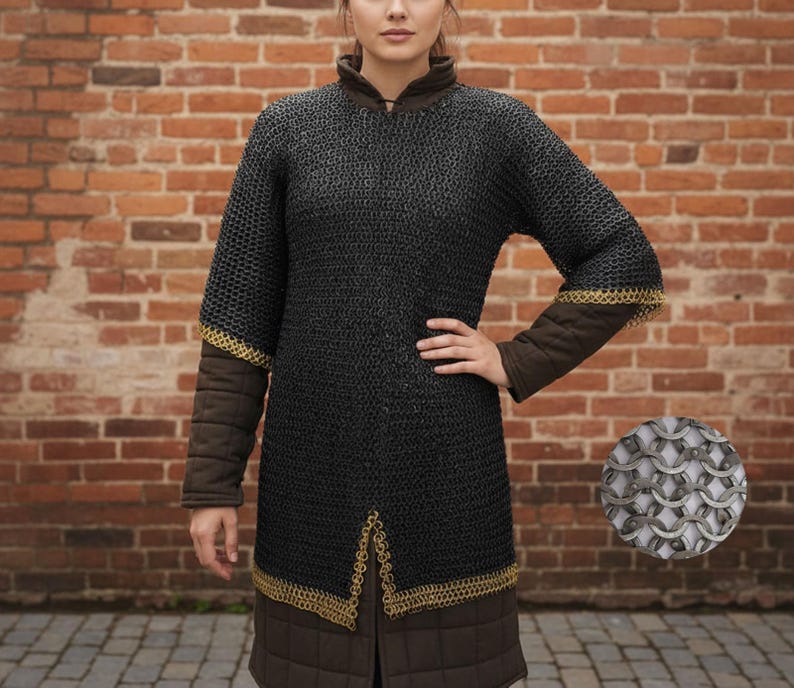 May include: Black chainmail shirt with gold trim on the sleeves and hem. The shirt has brown padded sleeves and a brown collar. A close-up of the chainmail is in the lower right corner. The background is a brick wall.