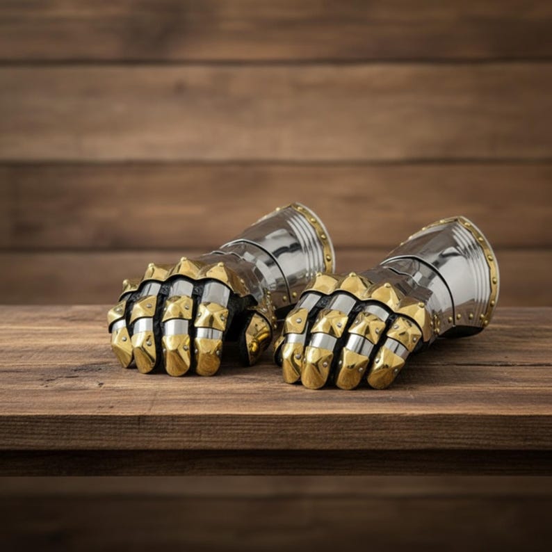 May include: A pair of medieval-style gauntlets, featuring a combination of silver and gold-colored metal. The gauntlets are designed with articulated finger plates and studded accents, resting on a wooden surface.