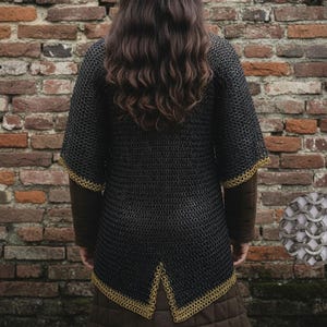 May include: Black chainmail shirt with gold trim on the sleeves and hem. The shirt is worn over a brown long-sleeved garment. The background is a weathered brick wall. A close-up of the chainmail is in the lower right corner.