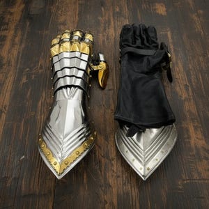 May include: A pair of medieval gauntlets. One is fully armored in silver metal with gold accents, the other is black leather with silver metal accents. The gauntlets are designed for protection and are displayed on a dark wood surface.