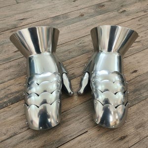 Pode incluir: Par de luvas de metal prateado polido. Cada luva apresenta um punho alargado, dedos articulados e placas sobrepostas para o pulso e a m&atilde;o. A superf&iacute;cie refletora do metal cria um efeito espelhado, refletindo o ambiente.