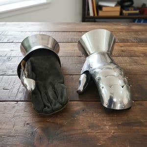 Pode incluir: Duas pe&ccedil;as de armadura medieval, uma manopla e uma luva, repousam sobre uma superf&iacute;cie de madeira. A manopla &eacute; feita de metal polido, com placas articuladas para os dedos e o pulso. A luva &eacute; de couro preto, projetada para caber na manopla.