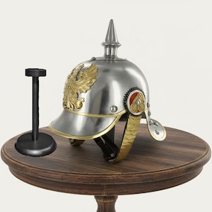 May include: A silver-colored Prussian Pickelhaube helmet with gold accents and an eagle emblem, displayed on a wooden table. A black helmet stand is to the left. The helmet has a prominent spike.