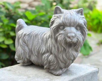 May include: A gray, detailed statue of a dog, likely a terrier, with textured fur and a focused expression. The sculpture is positioned on a stone surface, with a blurred green background of foliage.