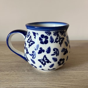 Może przedstawiać: Biały ceramiczny kubek z niebieskim rantem i uchwytem, ozdobiony powtarzającym się wzorem niebieskich motyli, kwiatów i liści. Kubek ma zaokrąglony kształt i wygodny uchwyt.