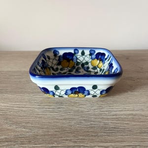 Może przedstawiać: Mała, kwadratowa ceramiczna miseczka z niebieską obwódką i kwiatowym wzorem. Wzór przedstawia niebieskie i żółte kwiaty z zielonymi liśćmi na białym tle. Miseczka jest prawdopodobnie używana do serwowania małych porcji.