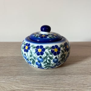 Könnte beinhalten: Eine runde Zuckerdose aus Keramik mit Deckel. Die Dose und der Deckel sind mit einem blauen Blumenmuster auf weißem Grund verziert. Der Deckel hat einen kleinen, runden Knopf. Die Dose steht auf einer Holzoberfläche.