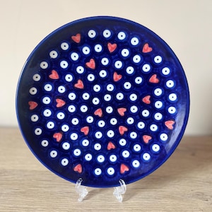 Puede incluir: Un plato redondo de cerámica azul oscuro con un patrón repetitivo de puntos blancos y corazones rojos. El plato se exhibe sobre un soporte de plástico transparente, mostrando su diseño decorativo. Un artículo de menaje.