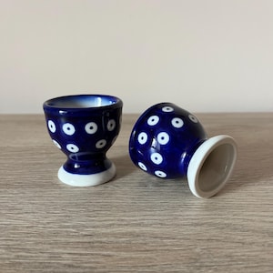 May include: Two ceramic egg cups. The egg cups are dark blue with white polka dots and a white base. One cup is upright, and the other is on its side. The egg cups are on a light brown wooden surface.