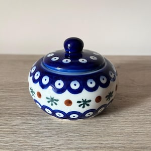 Könnte beinhalten: Runde Zuckerdose aus Keramik mit Deckel. Die Dose und der Deckel sind überwiegend blau und weiß, mit einem sich wiederholenden Muster aus weißen Punkten und blauen Kreisen. Die Dose hat auch braune und grüne Blumenakzente.