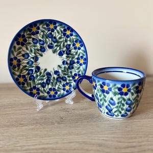 Op de afbeelding: Een witte keramische kop en schotel set. De kop en schotel zijn versierd met een blauw bloemmotief met gele centra en groene bladeren. De randen zijn blauw afgewerkt.