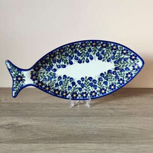 Könnte beinhalten: Eine keramische Fisch-Servierplatte mit blauem Blumendesign. Die Platte hat einen weißen Innenraum mit einem Rand aus blauen Blumen und grünen Blättern. Die Ränder sind blau eingefasst. Die Platte wird auf einem transparenten Ständer präsentiert.