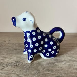 Op de afbeelding: Een keramieken beeldje van een gestileerde hond, met een witte kop en poten, en een donkerblauwe body versierd met witte stippen. De hond heeft een kleine, gebogen staart en een handvat-achtig kenmerk op zijn rug. Het beeldje staat tegen een lichte achtergrond.