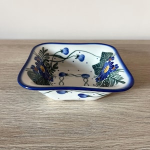 Może przedstawiać: Kwadratowy ceramiczny talerz z białą podstawą i niebieską obwódką. Talerz jest ozdobiony niebieskimi wzorami kwiatowymi i zielonymi liśćmi. Wzór zawiera niebieskie kwiaty i małe niebieskie jagody. Talerz stoi na drewnianej powierzchni.