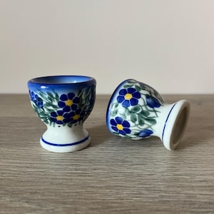 Op de afbeelding: Twee keramische eierdopjes met een bloemmotief. De bekers zijn wit met blauwe en gele bloemen en groene bladeren. De randen zijn blauw en de bodems hebben een blauwe streep.