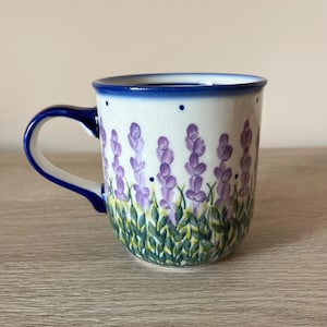 Peut inclure: Mug en céramique blanche avec une anse et un bord bleus, décoré de fleurs de lavande violettes et de feuilles vertes. La tasse a une forme arrondie et convient aux boissons chaudes. Le motif est peint à la main.