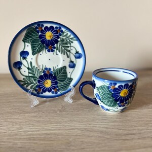 May include: A ceramic teacup and saucer set. The cup and saucer are white with a blue floral design. The flowers have blue petals and yellow centers, with green leaves. The rim of the cup and saucer are blue.