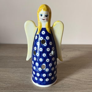 Può includere: Statuetta di angelo in ceramica con testa e ali gialle e un vestito blu decorato con pois bianchi. L'angelo ha un volto sorridente ed è progettato per contenere una candela. La statuetta è su una superficie di legno.