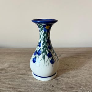 May include: A small, white ceramic vase with a flared top and a rounded body. The vase is decorated with blue floral patterns and green leaves. The rim and base have a blue trim. The vase is approximately 5 inches tall.