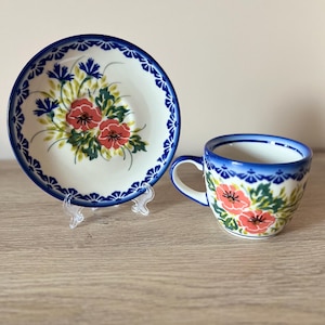 Op de afbeelding: Een keramische kop en schotel set. De witte stukken hebben een blauwe bloemenrand en een centraal ontwerp van rode bloemen, groene bladeren en blauwe accenten. De kop heeft een oor en de schotel is rond.