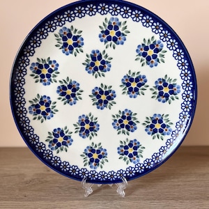 Könnte beinhalten: Ein runder Keramikteller mit weißer Basis, der ein sich wiederholendes Muster aus blauen Blumenclustern und einen blauen Rand aufweist. Der Rand ist mit einem zarten weißen und blauen Blumendesign verziert. Der Teller wird auf einem transparenten Ständer präsentiert.