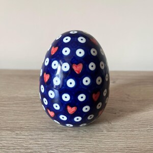 Pode incluir: Um ovo de cerâmica com um acabamento azul escuro brilhante. O ovo é decorado com um padrão de bolinhas brancas e pequenos corações vermelhos. O ovo decorativo é um item de decoração para casa único.