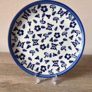 May include: A white ceramic plate with a blue rim and a repeating pattern of blue butterflies, flowers, and leaves. The plate is displayed on a clear plastic stand, showcasing its decorative design.