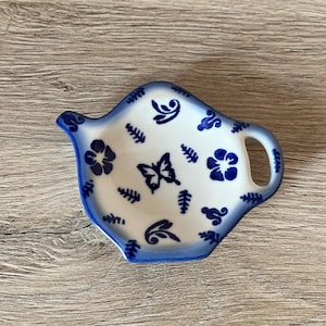 Könnte beinhalten: Ein weiß-blauer Keramik-Teebeutelhalter in Teekannenform. Er hat blaue Blumen- und Schmetterlingsmuster. Der Rand und der Griff sind blau. Der Teebeutelhalter ist etwa 13 cm lang und 10 cm breit.