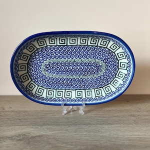 Könnte beinhalten: Eine ovale Servierplatte aus Keramik mit blauem Rand und komplizierten blauen und grünen Wirbel- und geometrischen Mustern. Die Platte wird auf einem transparenten Kunststoffständer präsentiert, der ihr dekoratives Design hervorhebt.