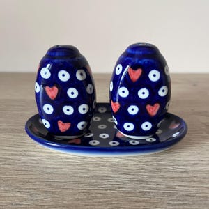 Könnte beinhalten: Ein Set aus zwei blauen Keramik-Salz- und Pfefferstreuern mit passendem Tablett. Die Streuer sind mit weißen Punkten und roten Herzen verziert. Das Tablett hat ein ähnliches Design. Das Set steht auf einer Holzoberfläche.