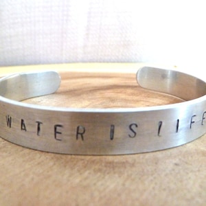 Pode incluir: Uma pulseira de metal prateado com as palavras "WATER IS LIFE" gravadas.