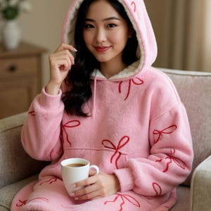 May include: A pink hooded blanket with red bow detailing. The blanket features a white fleece lining in the hood and a front pocket. A person is holding a white mug with a hot drink.