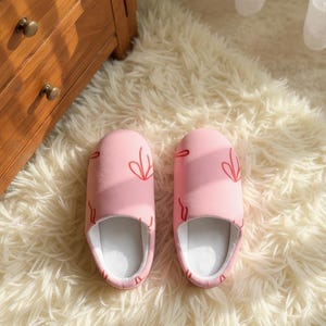 Peut inclure: Une paire de pantoufles roses avec un motif de feuille rouge. Les pantoufles ont une texture douce et moelleuse et un intérieur blanc. Elles sont conçues pour une utilisation en intérieur et sont posées sur un tapis blanc et duveteux.