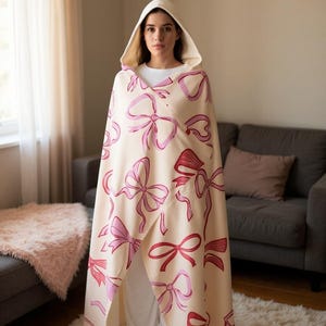 May include: A cream-coloured hooded cape featuring a pattern of pink and red bows. The cape is worn over a white outfit. The background includes a sofa, a window, and a white rug. The cape is a cozy garment.