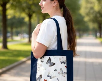 May include: A navy blue tote bag with a white panel displaying a butterfly design. The butterflies are in grey, black, and white, with red accents. The bag is carried over the shoulder, with a person in a white top.
