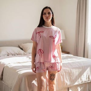 May include: A light pink pajama set featuring a short-sleeved top and matching shorts. The set is adorned with a repeating red bow design. The top has a white collar. The model is standing in front of a bed.