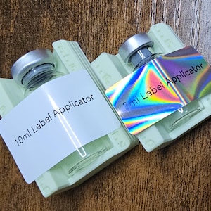 May include: Two small glass bottles with silver caps, each labeled as a "Label Applicator." One label reads "10ml," the other "3ml." The 3ml label has a holographic finish. Both bottles rest on light green holders.