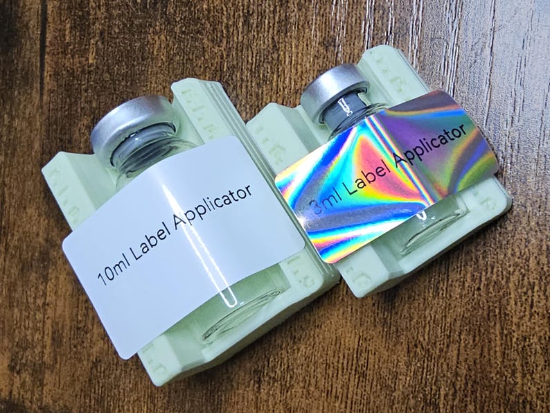 May include: Two small glass bottles with silver caps, each labeled as a "Label Applicator." One label reads "10ml," the other "3ml." The 3ml label has a holographic finish. Both bottles rest on light green holders.