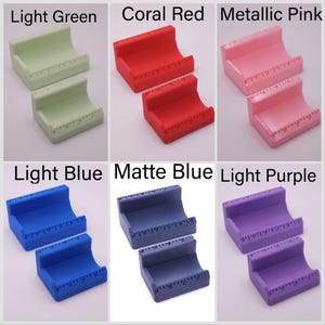 May include: A collection of six pairs of small, curved, plastic stands in various colors: light green, coral red, metallic pink, light blue, matte blue, and light purple. Each stand has a ruler-like marking along the top edge.