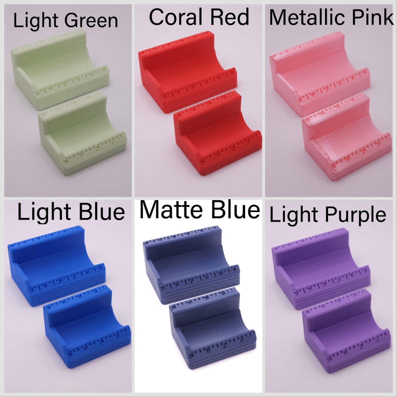 May include: A collection of six pairs of small, curved, plastic stands in various colors: light green, coral red, metallic pink, light blue, matte blue, and light purple. Each stand has a ruler-like marking along the top edge.