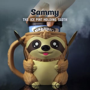 May include: A brown sloth-shaped holder designed to contain a pint of Ben & Jerry's ice cream. The sloth features a light tan belly, dark brown eye patches, and a small pink tongue. Two gold-coloured spoons are tucked into the sloth's arms.