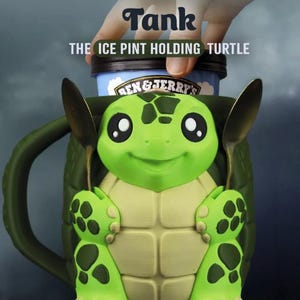 May include: A green turtle-shaped mug with a handle, featuring a light green plastron and dark green shell. The mug holds a Ben & Jerry's ice cream pint. Text above reads: Tank THE ICE PINT HOLDING TURTLE. Two metal spoons are tucked into the turtle's hands.