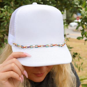 May include: A solid white trucker hat featuring a structured front panel and a mesh back. A decorative chain with multicolored enamel links in shades of blue, red, pink, and gold is attached across the base of the front crown above the brim.