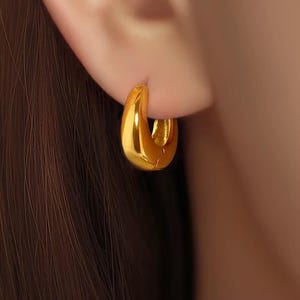 May include: A thick, polished gold hoop earring with a smooth, rounded surface and a high-shine metallic finish. The earring features a curved, chunky design that sits securely against the earlobe, reflecting light across its curved, reflective surface.